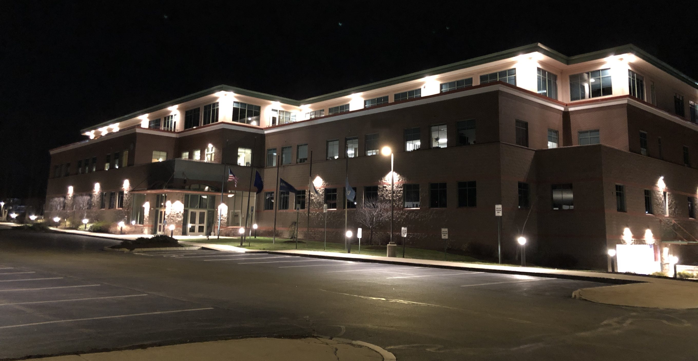 Office building at 75 New Hampshire Avenue lit at night by soffit downlights and wall-mounted lights, with a pole light at the entrance, Pease Tradeport, Portsmouth, NH